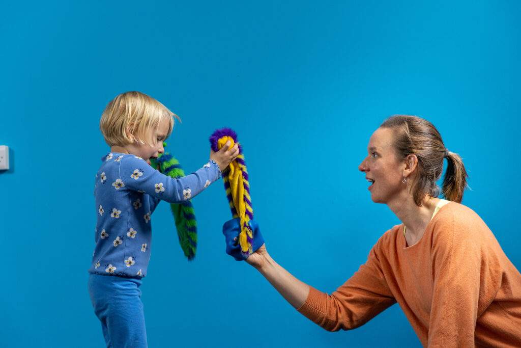 Against blue background small child on left in blue clothes and with blonde hair, holding a fluffy colourful caterpillar puppet in each hand. Right hand puppet hangs in the air and its tail is bitten by a blue rubber shark hand puppet operated by a woman in a peach coloured jumper opposite the girl. She looks at the girl's face and is on her eye level. The little girl instructed the play therapist how to play with the shark puppet.