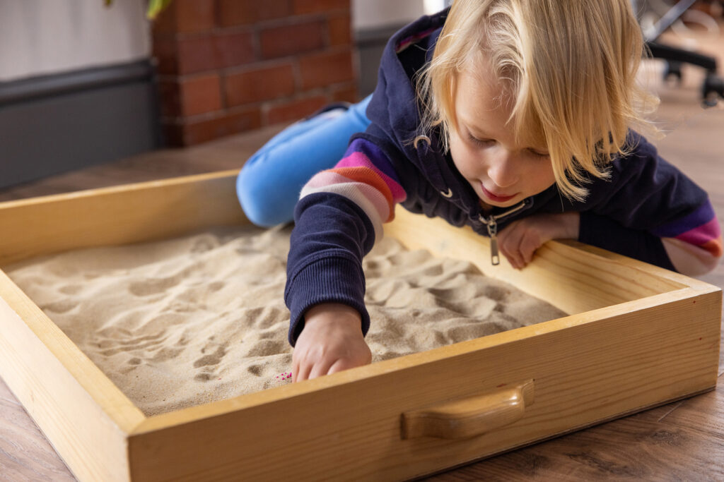 Young girl crawling into sand tray on a floor, looking at her right hand in the sand.